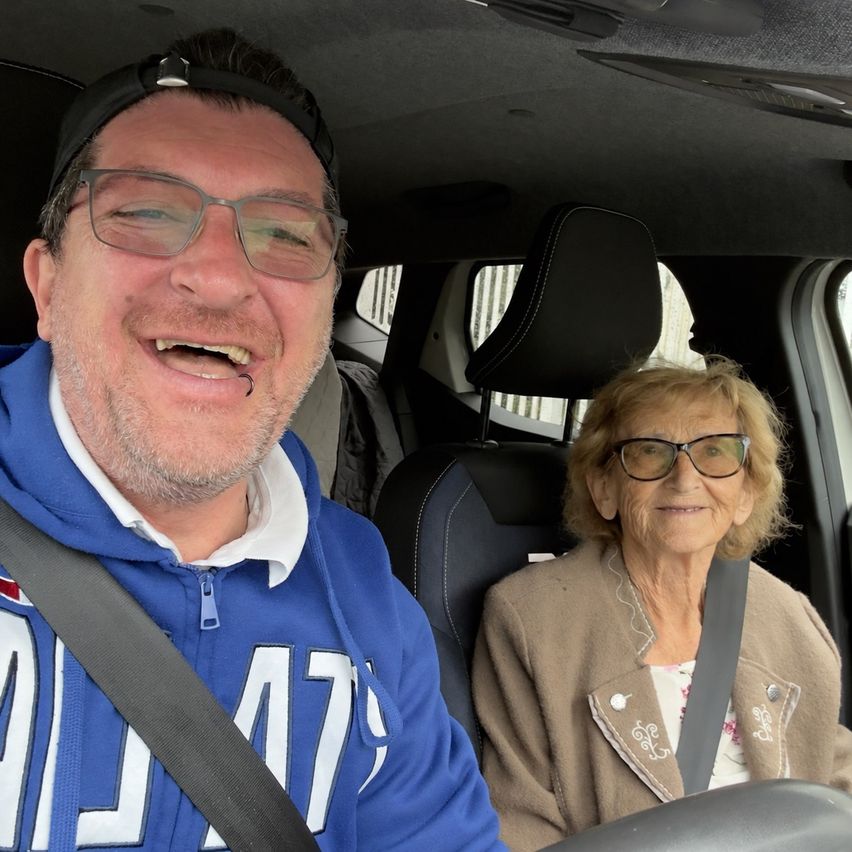A man wearing a blue hoodie with the number 47 and a woman wearing glasses sit in a car. The man is smiling and the woman is looking at the camera.