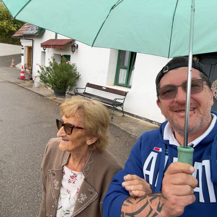 An elderly woman and a man are standing outside a house, holding a green umbrella. The woman is wearing sunglasses and a floral shirt, while the man has a tattoo on his arm. Behind them, there is a bench and a potted plant.