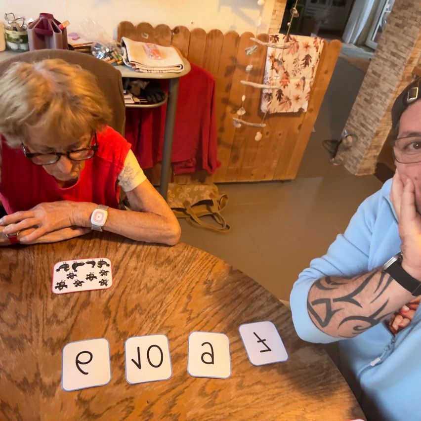An elderly woman wearing glasses and a red shirt is sitting at a table with a card game, while a man with tattoos looks on.
