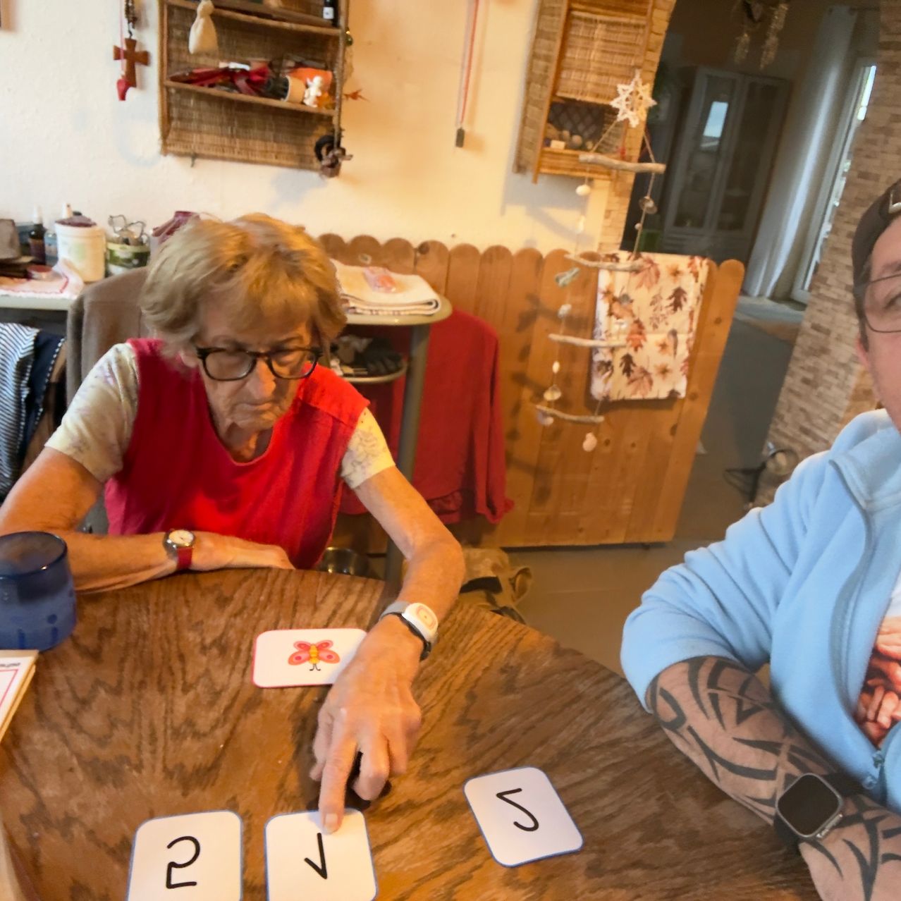 An elderly woman wearing glasses and a red vest is seated at a wooden table, playing a card game with a man wearing a blue shirt and a watch.