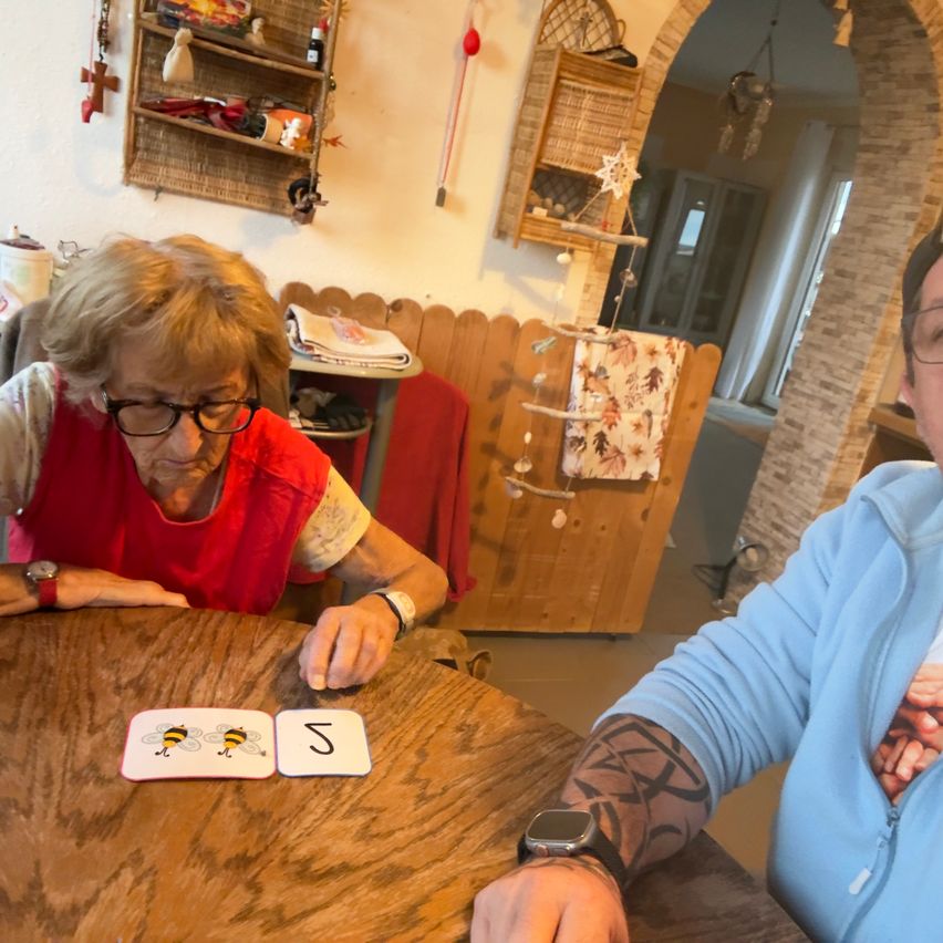 An elderly woman wearing glasses is sitting at a table with cards, while a man with a tattoo on his arm looks on.