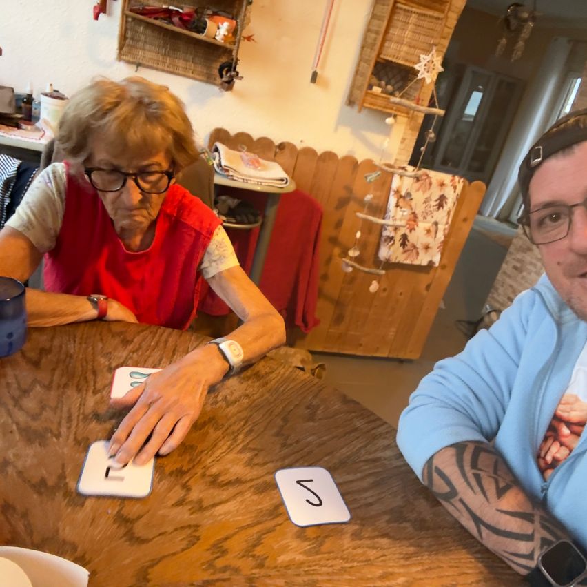 An elderly woman wearing glasses and a red vest plays a card game with a man in a blue shirt. They are seated at a wooden table with cards numbered 1 and 5.