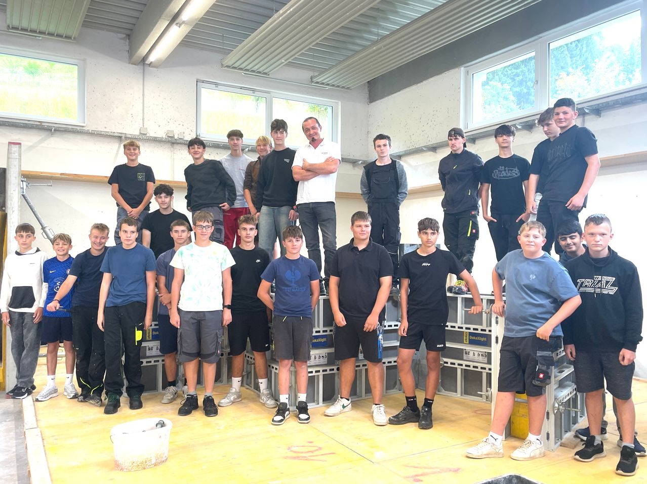 Eine Gruppe junger Männer, wahrscheinlich Schüler, steht auf Kisten in einem Raum. Einige sind in Freizeitkleidung und ein Mann in einem weißen Hemd steht mit verschränkten Armen oben auf den Kisten.