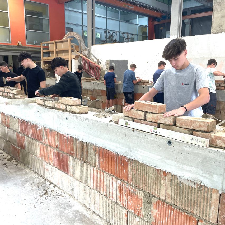 Eine Gruppe junger Männer arbeitet an einer Ziegelmauer, möglicherweise an einer Baustelle. Sie tragen Freizeitkleidung und einige haben eine Brille. Sie stehen auf der Ziegelmauer und einige sind am Boden. Eine Wasserwaage ist auf der Mauer platziert.