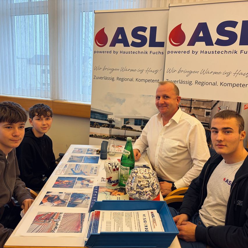 Vier Personen sitzen lächelnd an einem Tisch mit Werbematerialien. Dahinter befinden sich Banner mit dem Text 'ASL powered by Haustechnik Fuchs'.