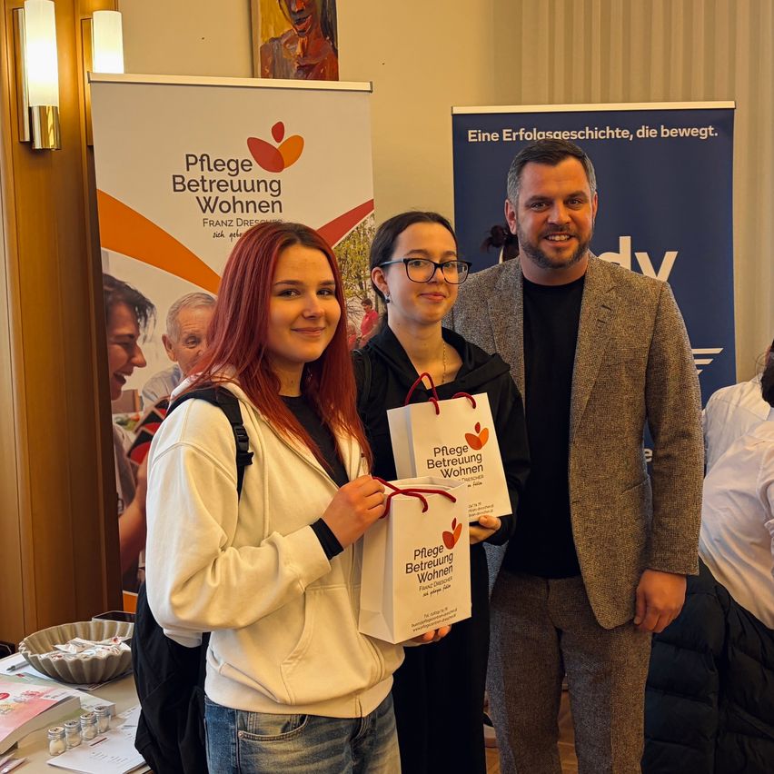 Drei Menschen stehen lächelnd zusammen. Eine Frau hält eine Geschenktüte mit 'Pflege Betreuung Wohnen' darauf. Dahinter steht ein Plakat mit den gleichen Worten und einem Bild von Menschen.