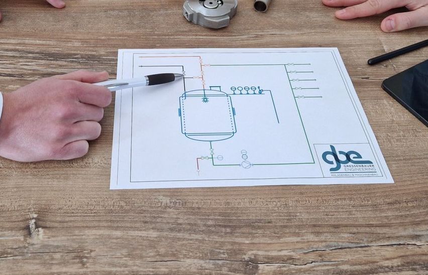 Zwei Hände halten einen Stift und zeichnen auf einen Plan mit Ventilen und Rohren auf einem Holztisch. Ein Tablet und ein Stift liegen ebenfalls auf dem Tisch.