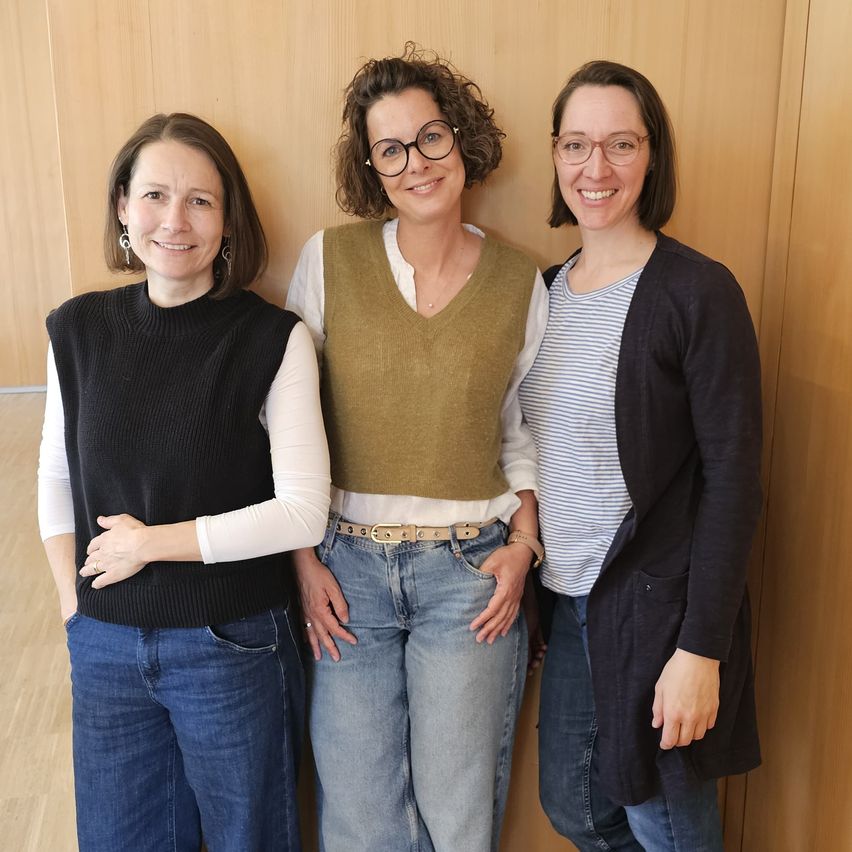 Drei Frauen stehen gemeinsam vor einer Holzwand, lächeln und posieren für ein Foto. Die Frau auf der linken Seite trägt ein schwarzes Rollkragenshirt, die Frau in der Mitte trägt ein grünes Weste und Jeans, und die Frau auf der rechten Seite trägt einen schwarzen Cardigan.