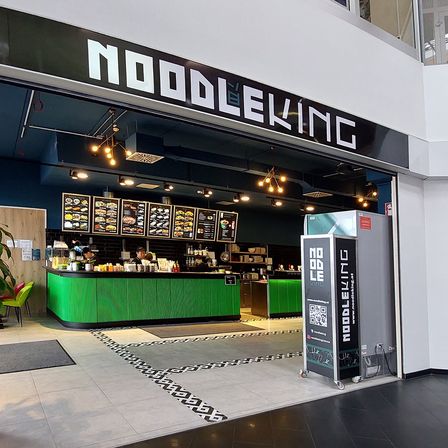 Ein Restauranteingang mit einem schwarz-weißen Schild, auf dem 'Nudelkönig' steht. Es gibt eine grüne Theke mit Essen und Getränken. Ein Menübrett mit Bildern von Speisen und Text.