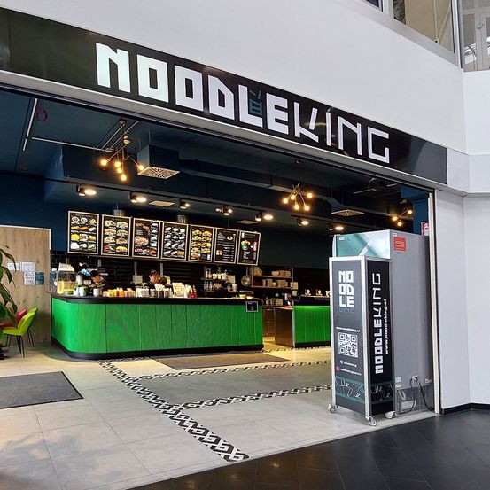 Ein Restauranteingang mit einem schwarz-weißen Schild, auf dem 'Nudelkönig' steht. Es gibt eine grüne Theke mit Essen und Getränken. Ein Menübrett mit Bildern von Speisen und Text.
