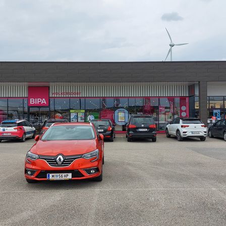 Ein roter Renault Clio ist vor dem BIPA-Geschäft geparkt. Andere Autos stehen auf dem Parkplatz. Hinter dem Geschäft befindet sich eine Windturbine.