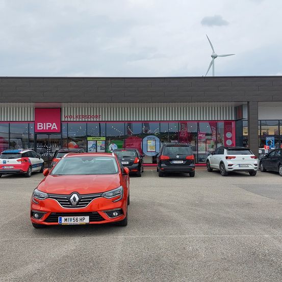 Ein roter Renault Clio ist vor dem BIPA-Geschäft geparkt. Andere Autos stehen auf dem Parkplatz. Hinter dem Geschäft befindet sich eine Windturbine.