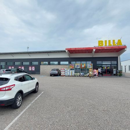 Außenansicht eines BILLA-Ladens mit einem großen gelben Schild, vor dem geparkte Autos und einige Personen, die in der Nähe des Eingangs gehen.