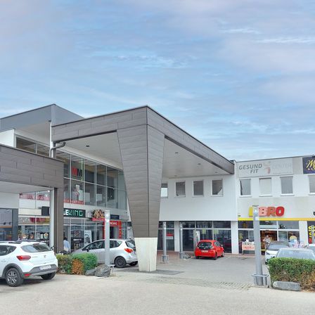 Außenansicht eines Einkaufszentrums mit mehreren Geschäften, darunter ein Fitnesscenter und ein Geschäft mit der Aufschrift 'BRO'. Vor dem Zentrum sind Autos geparkt, und eine Person steht in der Nähe des Eingangs.