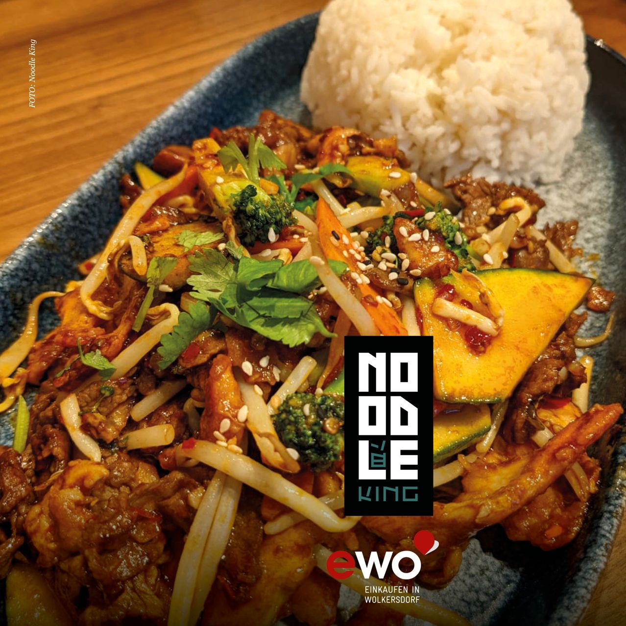 Ein Teller mit asiatischem Stir-Fry mit Nudeln, Gemüse und Fleisch. Eine Reiskugel befindet sich auf der Seite. Der Teller steht auf einer Holzoberfläche.