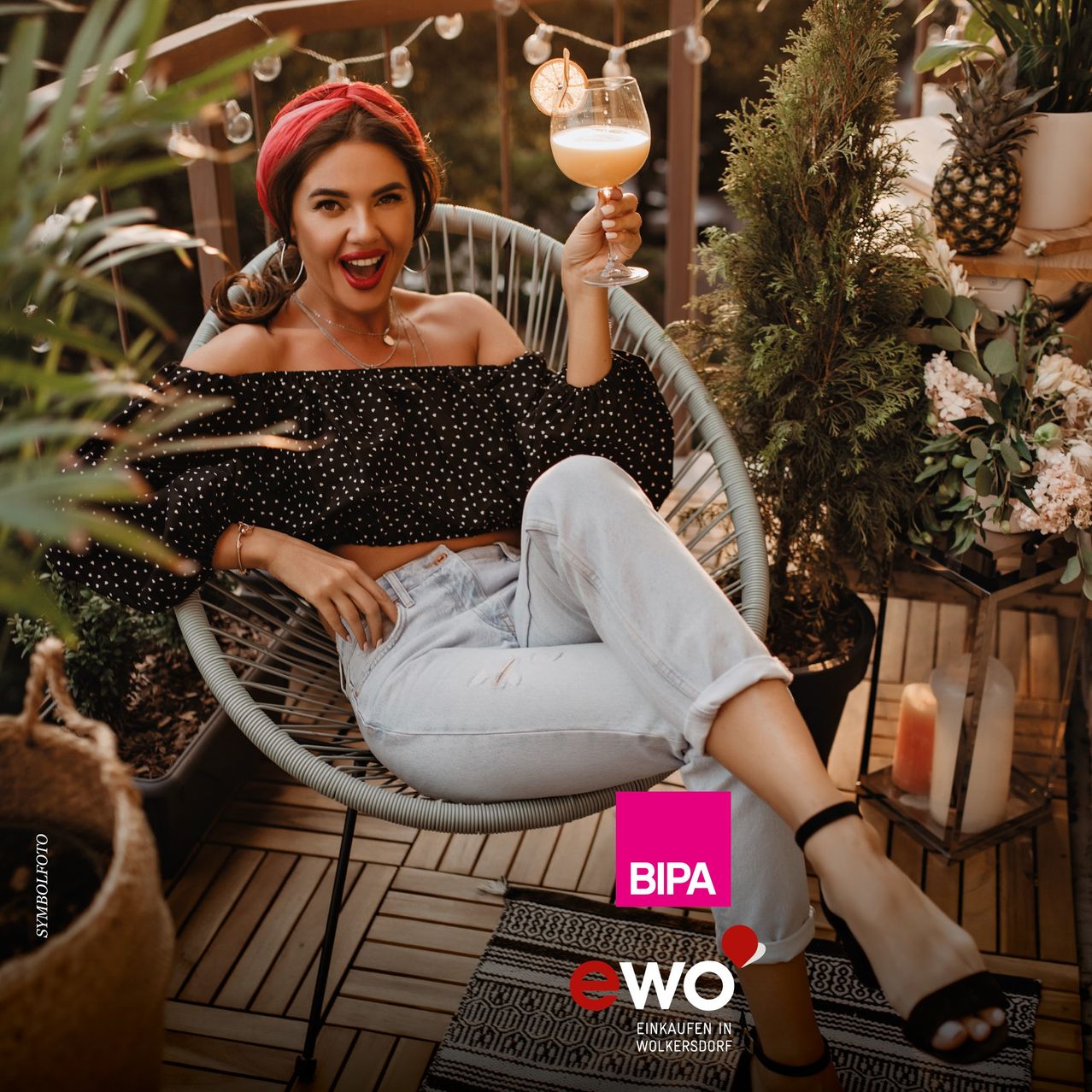 Eine Frau in einem gepunkteten Oberteil und Jeans sitzt auf einem Stuhl, hält ein Glas Wein in der Hand und lächelt mit den Zähnen. Sie befindet sich auf einem Balkon mit Pflanzen und einem Holzboden.