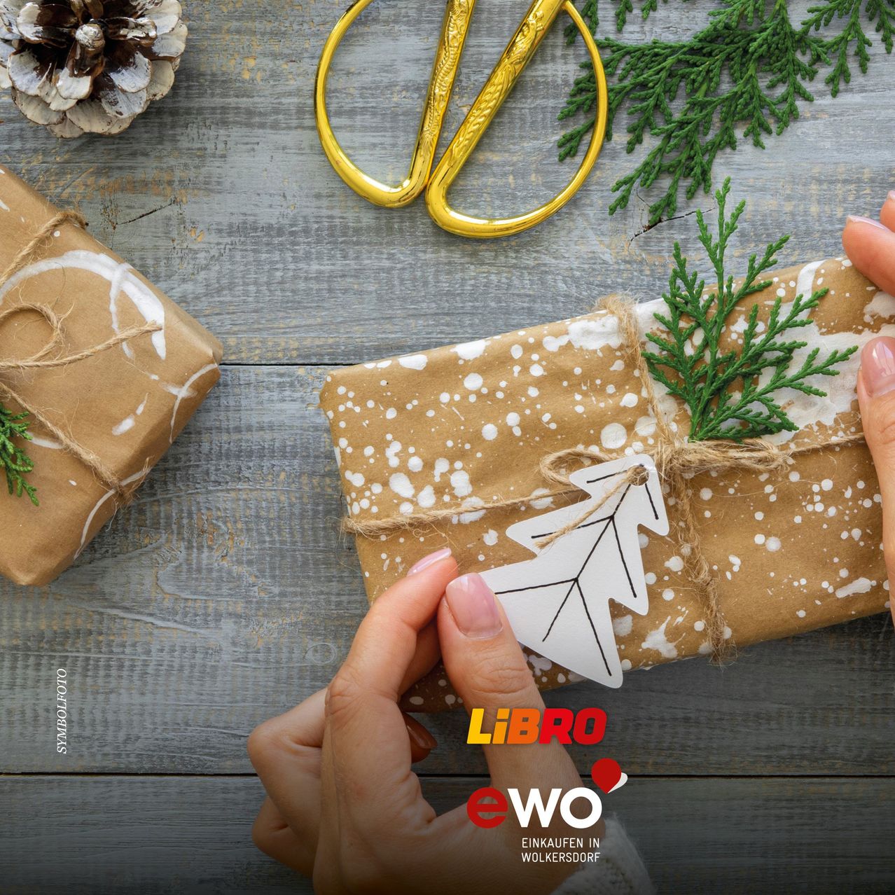 Eine Person wickelt ein Geschenk mit braunem Papier und einem weißen Anhänger in Form eines Weihnachtsbaums ein. Das Geschenk liegt auf einem Holztisch mit einer goldenen Schere und einer Tannenzapfen.