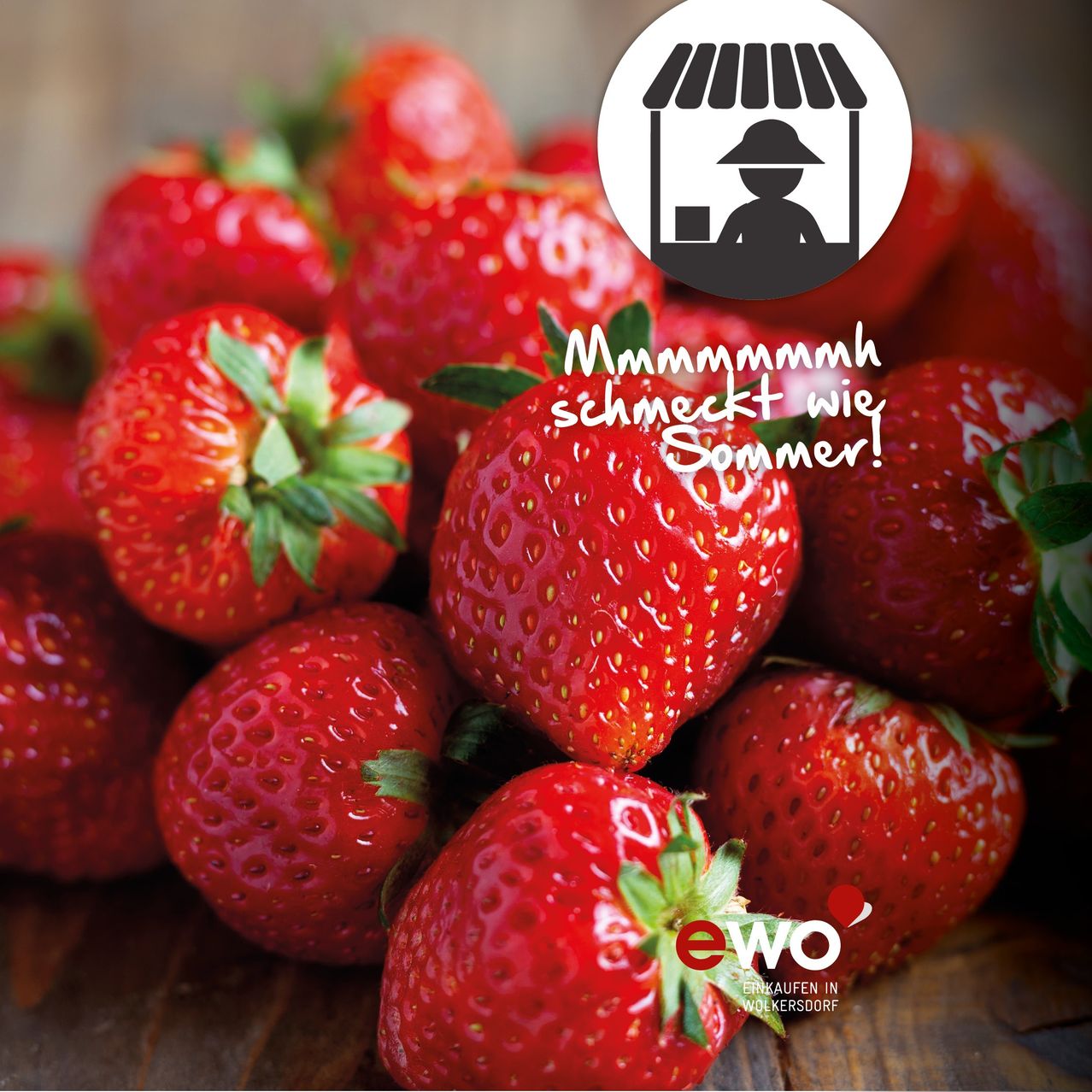 Ein Stapel Erdbeeren auf einem Holztisch. Das Bild zeigt ein weißes kreisförmiges Logo mit einem Häuschen und einer Person sowie den Text 'Mmmm schmeckt wie Sommer!' in großen Buchstaben.