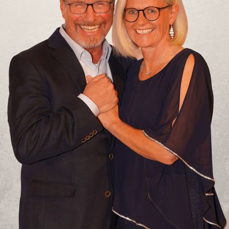 Ein Mann und eine Frau stehen eng beieinander und lächeln. Der Mann trägt einen schwarzen Anzug und eine Brille. Die Frau trägt ein schwarzes Kleid mit silbernen Borten und Ohrringe. Sie könnten für ein Foto posieren.