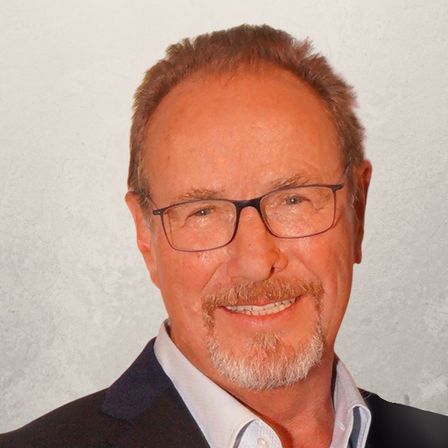 Ein Porträt eines älteren Mannes mit Bart, der einen schwarzen Anzug und eine Brille trägt, lächelt vor einem hellen Hintergrund.