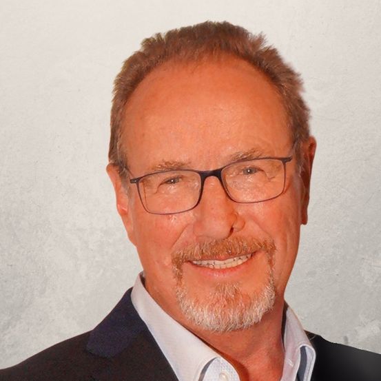 Ein Porträt eines älteren Mannes mit Bart, der einen schwarzen Anzug und eine Brille trägt, lächelt vor einem hellen Hintergrund.