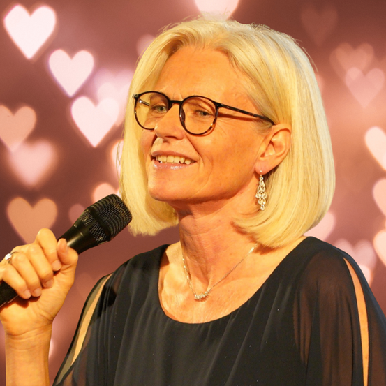 Eine Frau mit kurzen blonden Haaren, die eine Brille und Ohrringe trägt, hält ein Mikrofon. Sie lächelt und scheint zu singen. Der Hintergrund ist rosa mit herzförmigen Schatten.