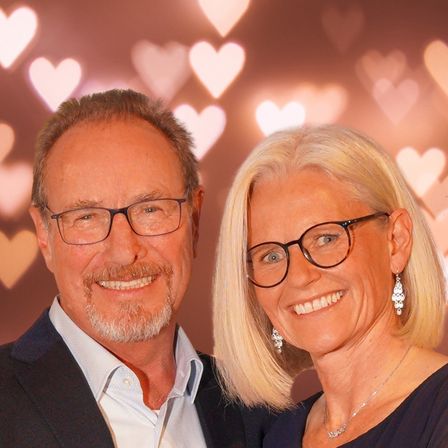 Ein Mann und eine Frau mit Brille lächeln warm in die Kamera, mit einem Hintergrund aus Herzformen.