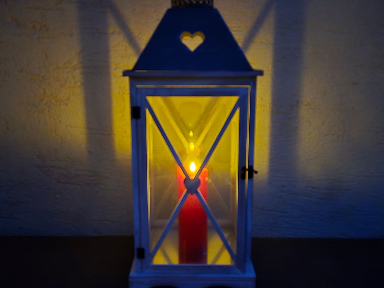 Eine blaue Laterne mit einem herzförmigen Design auf der Oberseite, beleuchtet von einer einzelnen roten Kerze im Inneren, die ein warmes Licht an eine Wand wirft.
