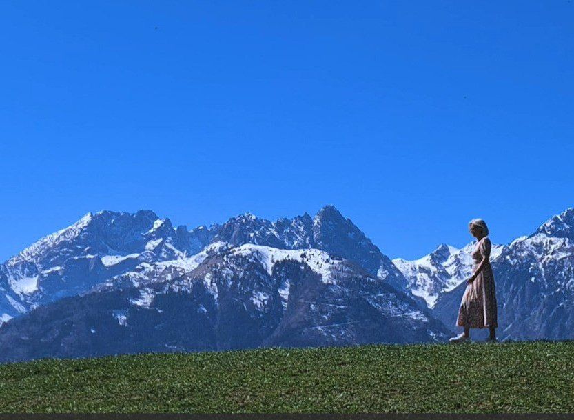 A woman stands on a grassy hill with snow-capped mountains in the background. The sky is clear and blue.