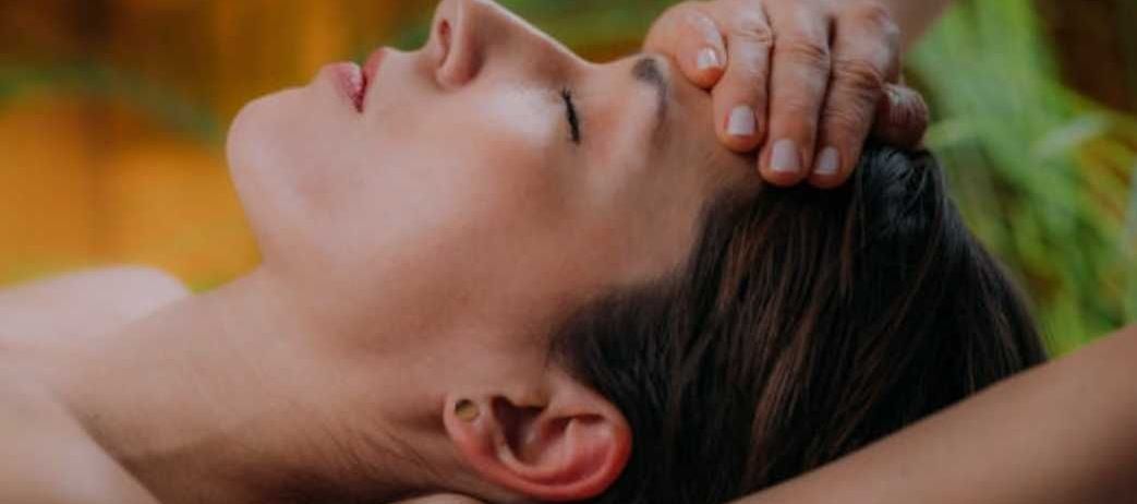 Eine Frau liegt auf dem Rücken und bekommt eine Kopfmassage von einer anderen Person. Die Masseuse drückt sanft mit ihren Händen auf den Kopf der Frau.