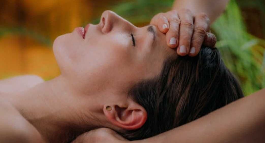 Eine Frau liegt auf dem Rücken und bekommt eine Kopfmassage von einer anderen Person. Die Masseuse drückt sanft mit ihren Händen auf den Kopf der Frau.