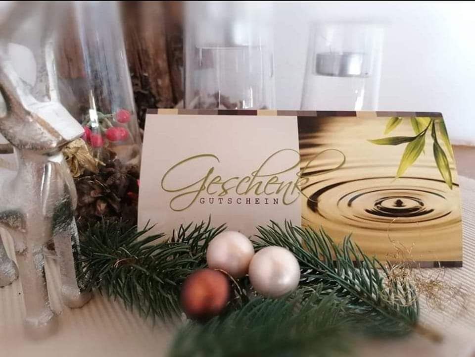 Eine Feiertagskarte mit dem Text Geschenk Gutschein, die eine goldene Wasserwelle und Kiefernzweige zeigt. Sie ist umgeben von einem roten Ornament, Tannenzapfen und zwei Glasbehältern.