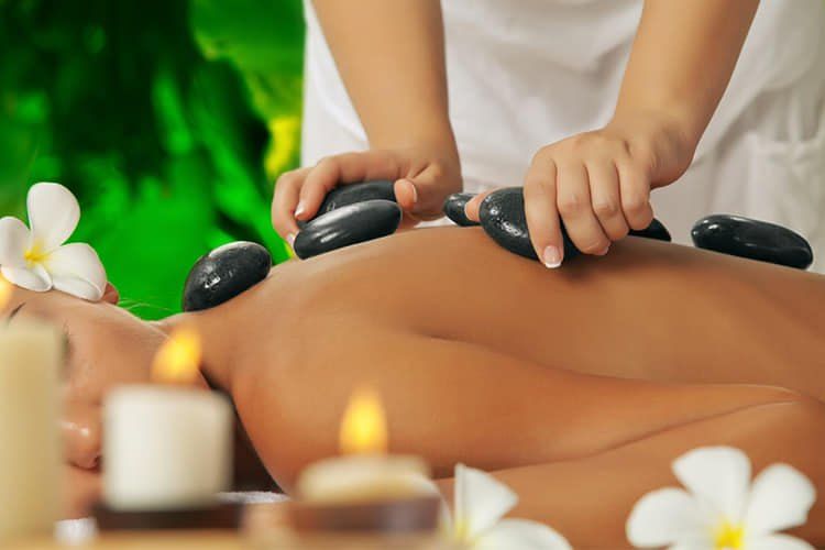 Eine Frau erhält in einem Spa eine Rückenmassage mit heißen Steinen. Sie liegt auf dem Bauch, mit Blumen und Kerzen um sie herum.