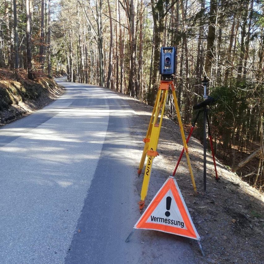 Ein Vermessungsinstrument steht auf einem Stativ am Straßenrand, mit einem Warnschild, das 'Vermessung' sagt. Die Straße ist von Bäumen umgeben, und der Boden ist mit kleinen Steinen bedeckt.