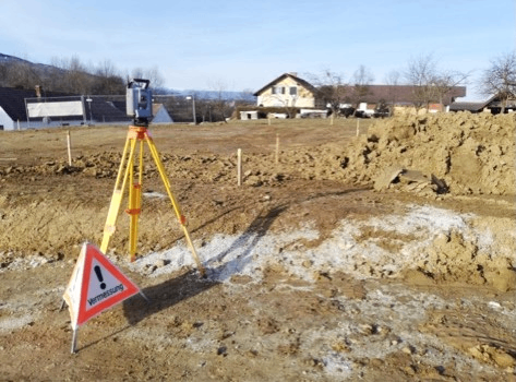 Ein Vermessungsinstrument steht auf einem Stativ auf einer Baustelle mit einem Warnschild in der Nähe, Häusern und Bäumen im Hintergrund.