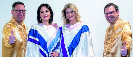 Four people in matching outfits pose for a photo, two women in the middle wearing long, flowing dresses, and two men on the sides wearing gold and white outfits. They all have happy expressions.