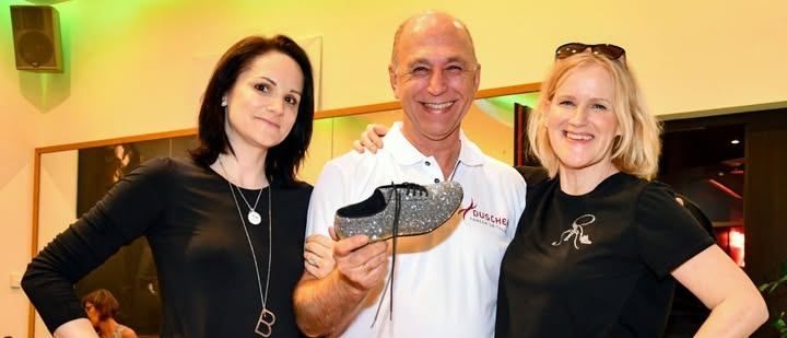 Drei Menschen posieren für ein Foto, ein Mann hält einen Schuh, zwei Frauen lächeln, eine trägt eine Uhr, in einem Raum mit Holzboden und einem Spiegel.