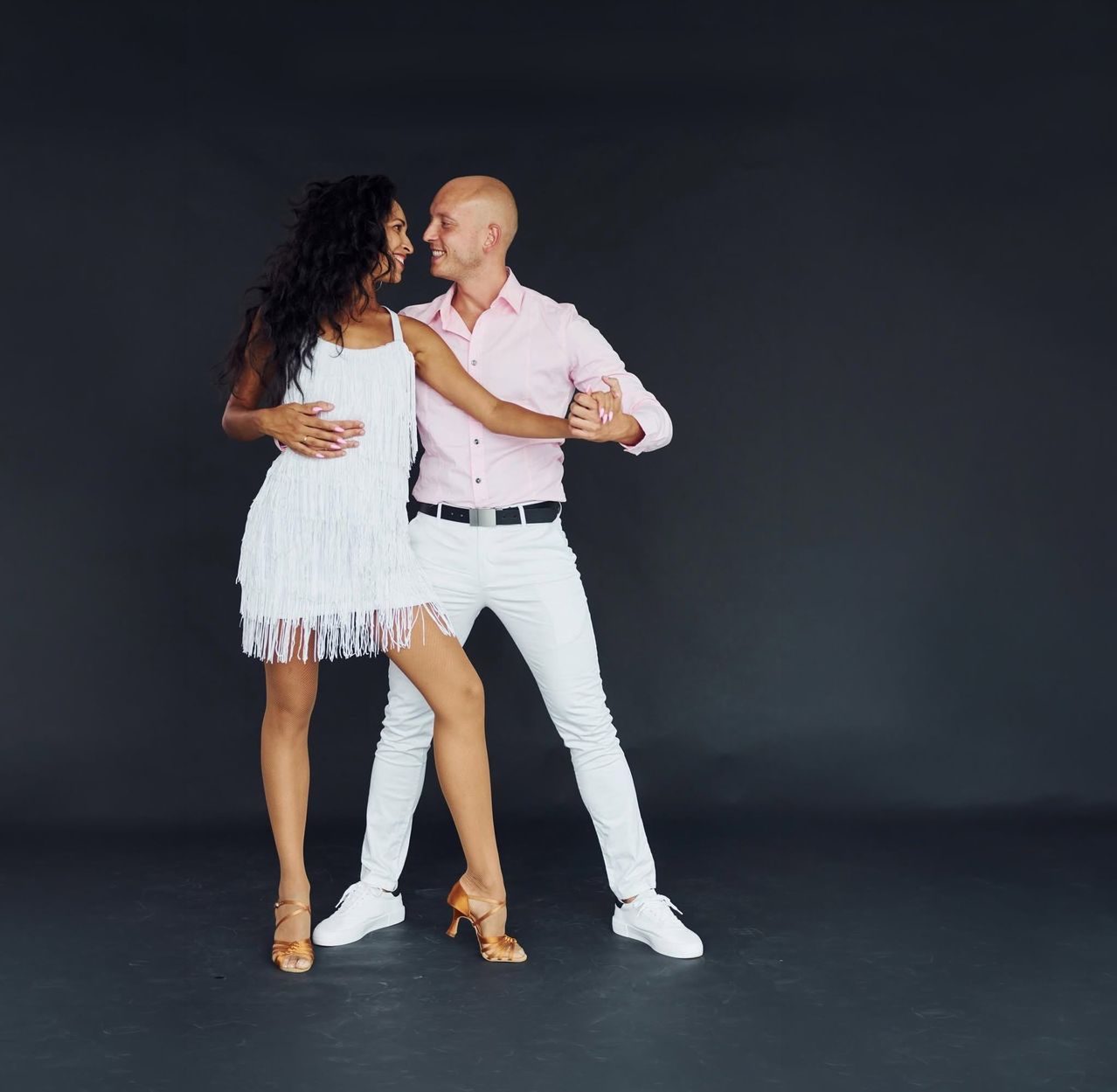 A man and woman are dancing together, smiling, and dressed in white and pink. The woman is wearing a white dress with fringes.