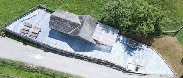 Luftaufnahme eines dreieckigen Hauses auf einem Hügel mit einer Schotterauffahrt, Holzbänken und einem eingezäunten Bereich. Bäume und Gras umgeben das Gebiet.