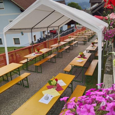 Ein Freiluft-Essbereich unter einem weißen Zelt, mit Reihen von Holztischen und Bänken, Blumen und Notizen auf den Tischen. Ein blaues Gebäude und ein rotes Auto sind im Hintergrund zu sehen.