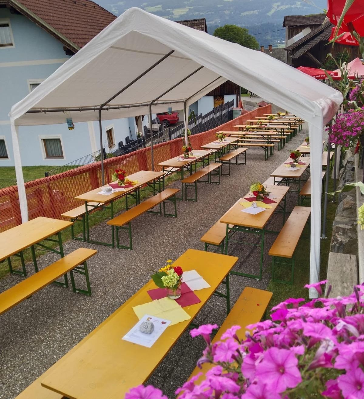 Ein Freiluft-Essbereich unter einem weißen Zelt, mit Reihen von Holztischen und Bänken, Blumen und Notizen auf den Tischen. Ein blaues Gebäude und ein rotes Auto sind im Hintergrund zu sehen.