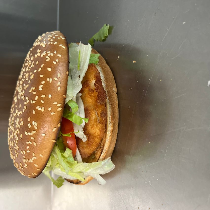 Ein halbgeschnittener Burger mit Sesambrötchen, Salat, Tomate und einer panierten Hühnchen-Patty auf einer Edelstahloberfläche.