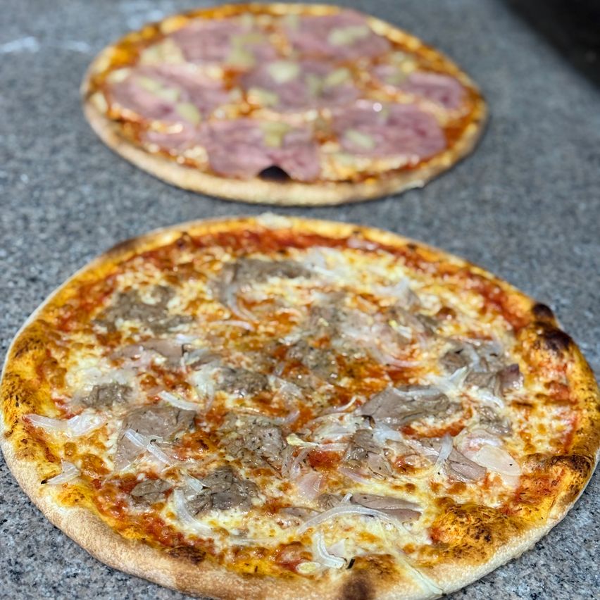 Zwei Pizzen auf einem Tisch. Eine hat eine rote Soße und rosa Beläge, die andere einen goldbraunen Rand mit einer Mischung aus Fleisch und Zwiebeln.