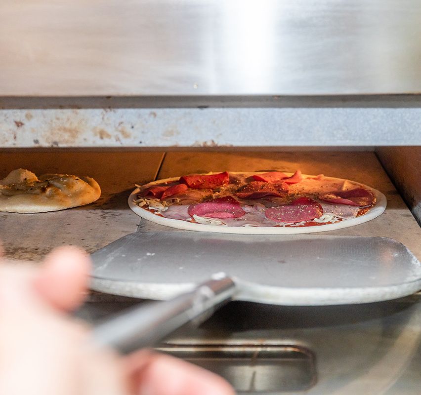 Eine Pizza wird mit einem Pizzaschaufel aus dem Ofen genommen. Eine andere Pizza ist auf dem Tresen.