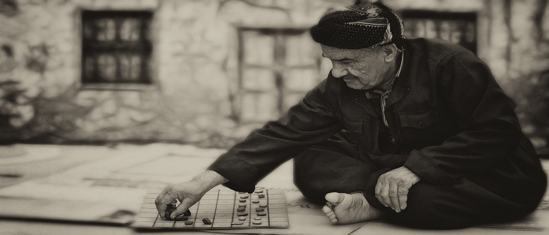 Ein älterer Mann sitzt barfuß auf dem Boden und spielt ein Spiel mit kleinen Steinen, entweder Schach oder Dame. Er ist konzentriert und ernst, trägt einen Hut und eine Jacke.