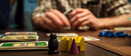 Eine Person sitzt an einem Tisch und spielt ein Brettspiel mit Karten und Holzteilen.