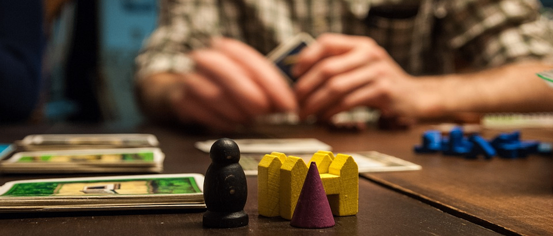 Eine Person sitzt an einem Tisch und spielt ein Brettspiel mit Karten und Holzteilen.