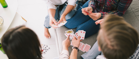 Mehrere Menschen sitzen auf einer Couch und spielen Karten. Einige Karten werden gehalten, andere liegen auf dem Tisch.