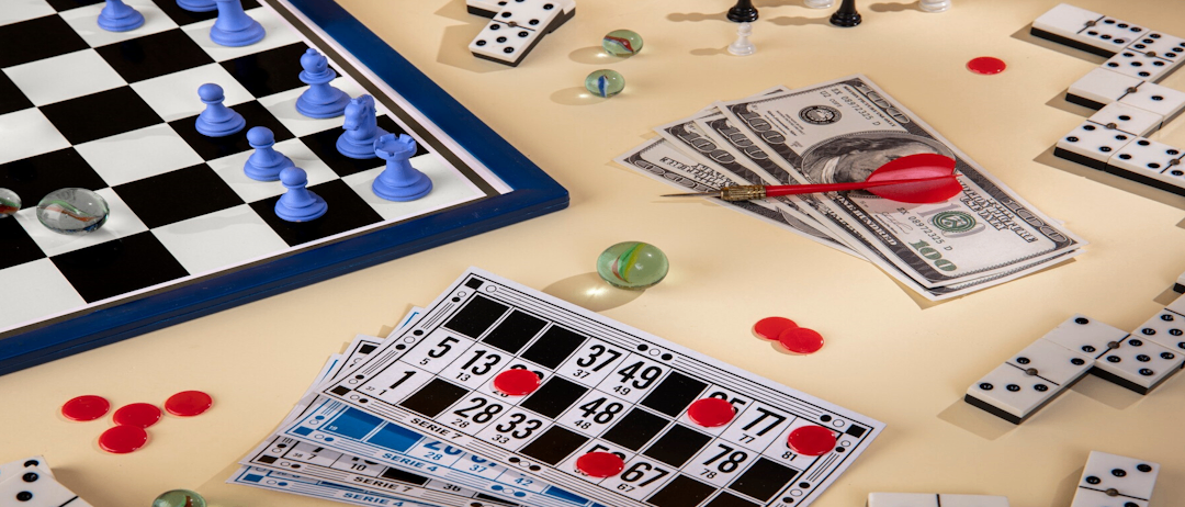 Auf einem Tisch liegt eine Schachtel, Würfel, Murmeln und ein Pfeil mit roter Spitze. Geld und eine Bingokarte mit roten Spielsteinen sind ebenfalls vorhanden.