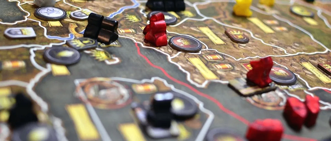 Ein Strategiebrettspiel ist auf einem Tisch aufgebaut, mit verschiedenen farbigen Spielsteinen, einschließlich rot, schwarz und gelb, auf einem kartenartigen Layout. Das Spiel beinhaltet runde und quadratische Spielsteine, und der Hintergrund ist eine Karte mit verschiedenen Regionen.